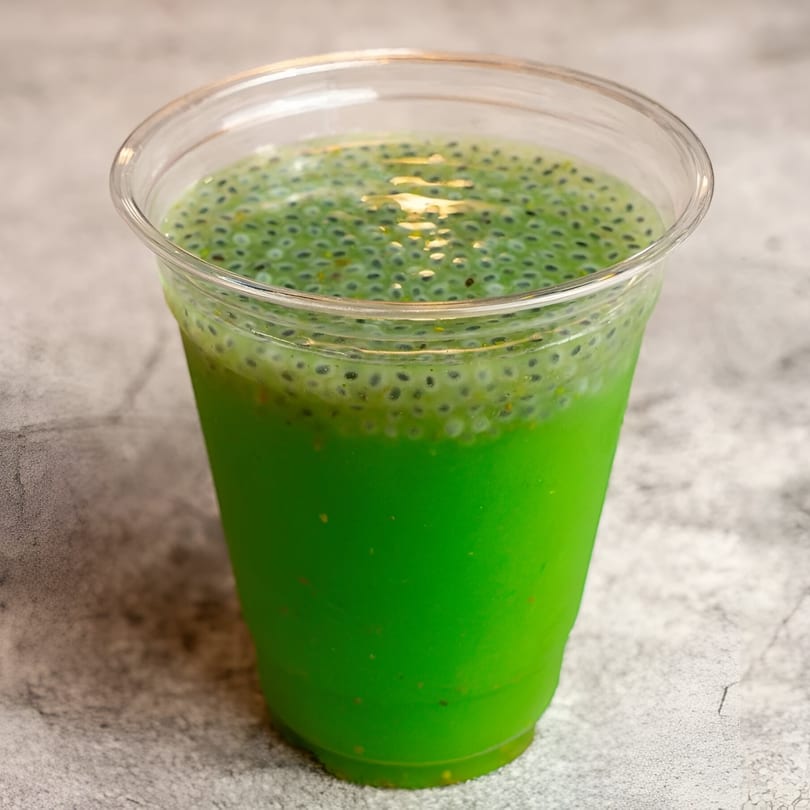 Bright green Indian drink with basil seeds in a clear plastic cup on a gray surface.