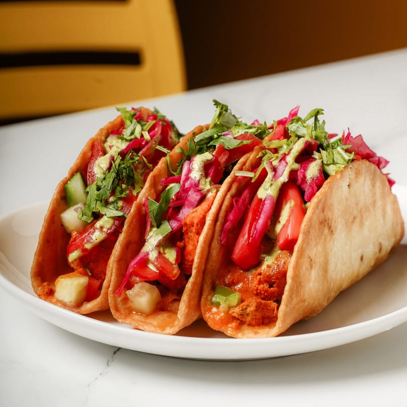 Three vibrant Indian tacos on a white plate, filled with chicken, red cabbage, and green sauce.